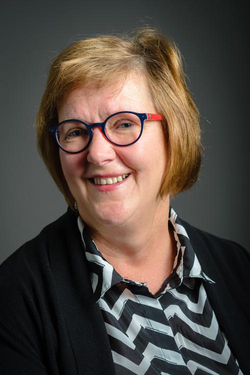 Muotokuva 64-vuotiaasta silmälasipäisestä hymyilevästä naisesta. Naisella on sinipunaiset pyöheähköt silmälasit, vaaleanpunertavat polkkapituiset hiukset, mustavalkokuvioinen paitapusero ja musta neuletakki.